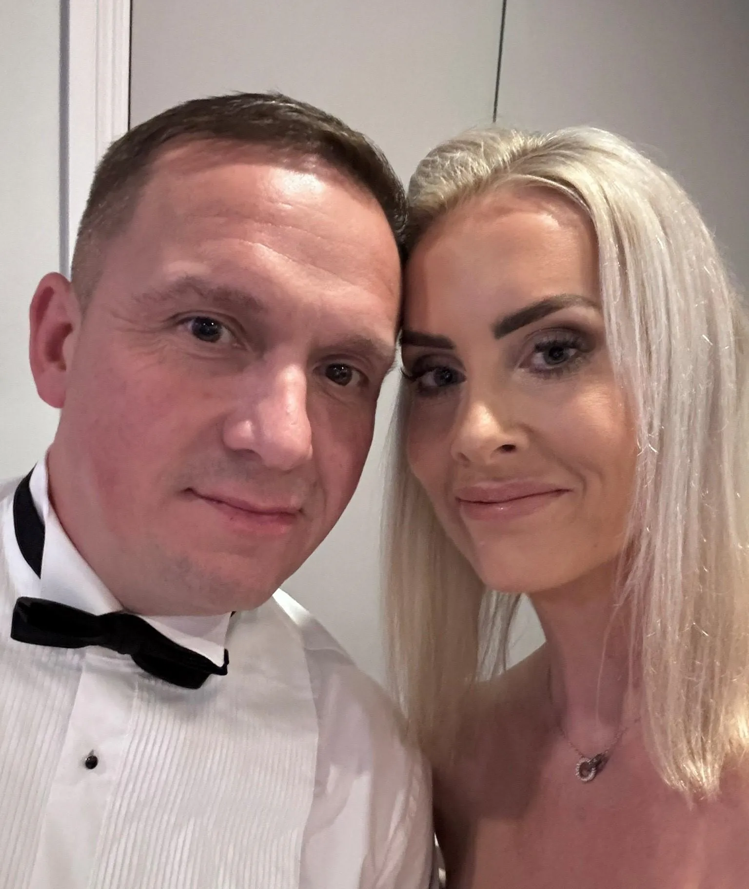 A smiling couple poses closely for a photo. The man wears a white tuxedo shirt with a black bow tie