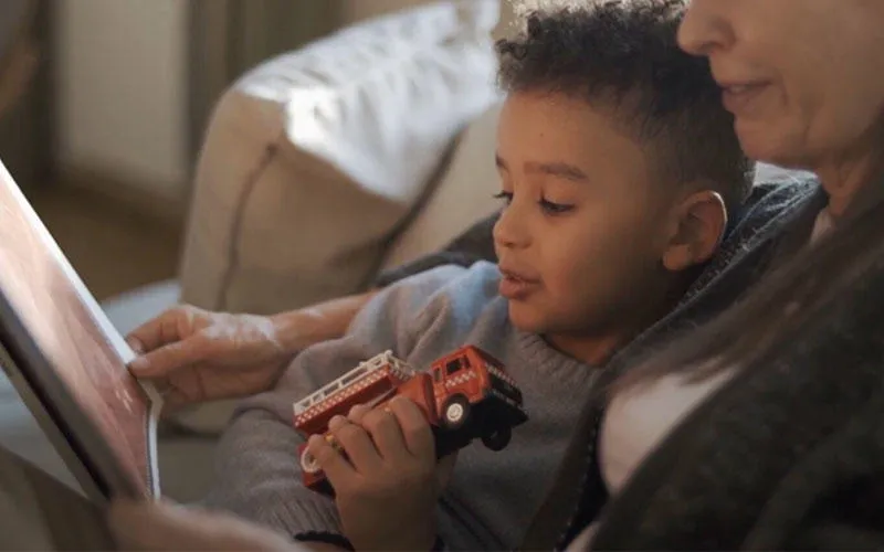 Young boy holding a toy truck sat reading with a woman 